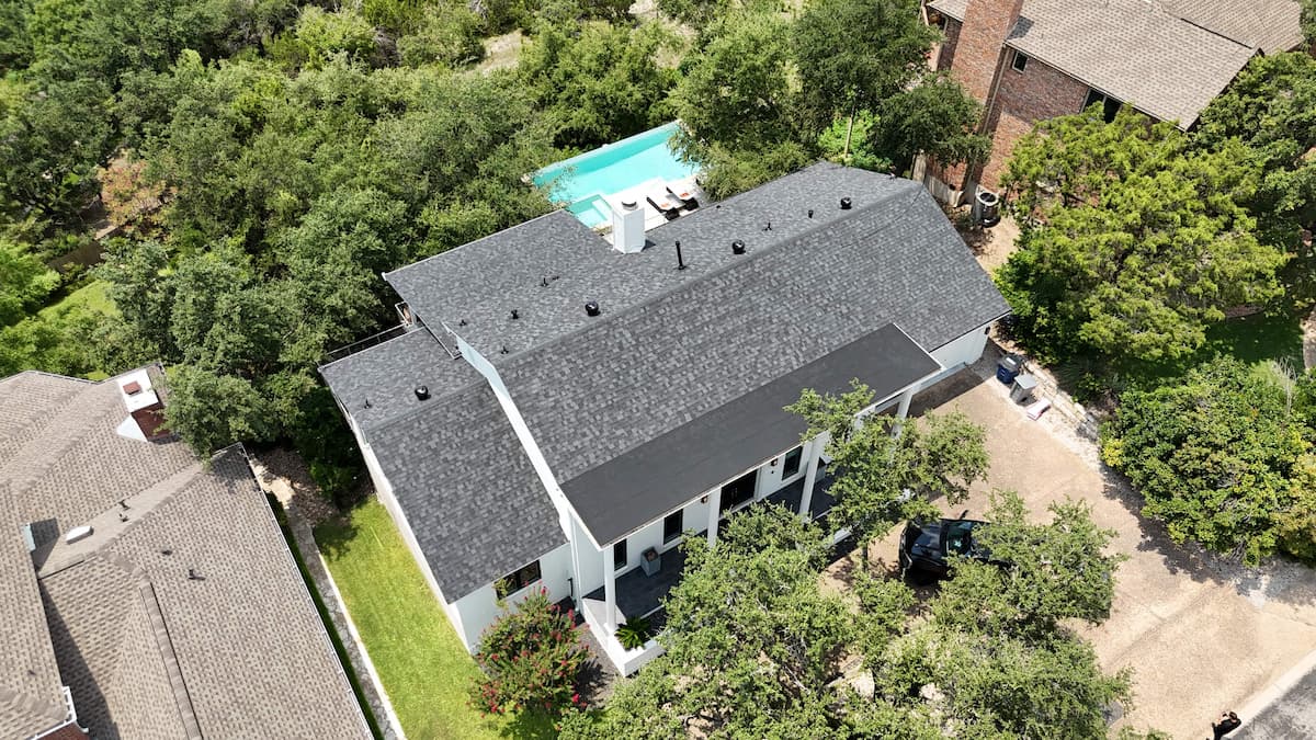 Aerial drone photo of a multi-level residential house with a newly replaced charcoal grey shingle roof, patio, and swimming pool surrounded by lush trees in Austin, TX.