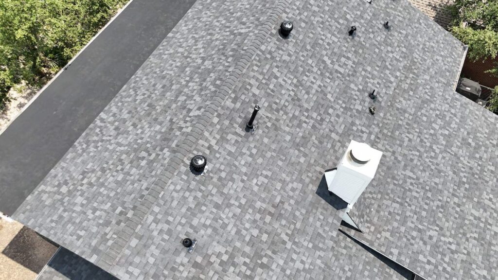Aerial drone photo showing a completed residential roof replacement featuring light grey architectural asphalt shingles, modified bitumen transition roofing, white chimney flashing, and attic vents in Austin, TX.