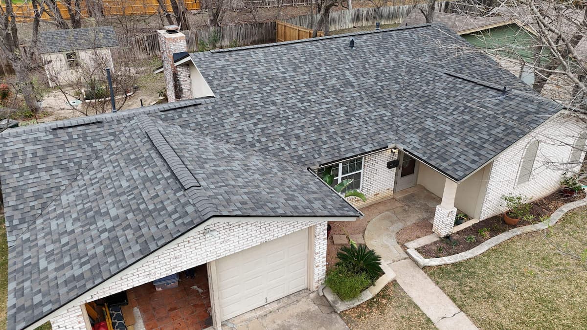 Elevated drone shot of a brick ranch-style home featuring a newly replaced Owens Corning Williamsburg Grey architectural shingle roof, stone chimney, and attached garage in Austin, TX.