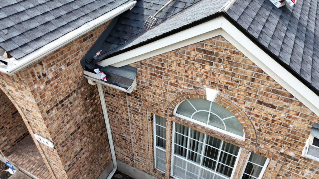 Brick home roof edge during tear-off with exposed underlayment above the gutter and dark drip edge along the rake edge